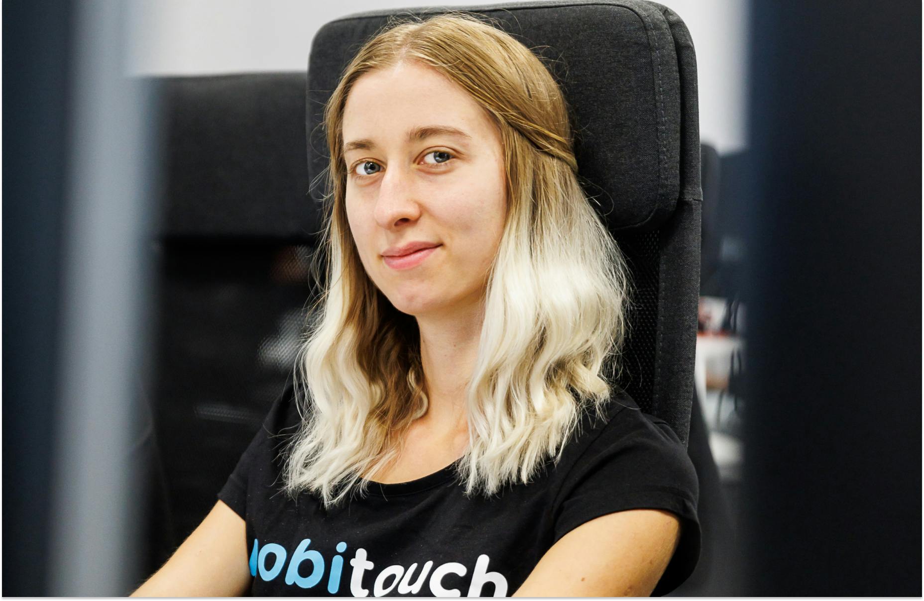 Kobieta, programistka, w blond włosach, z lekkim uśmiechem na twarzy, siedząca na krześle przez monitorem
