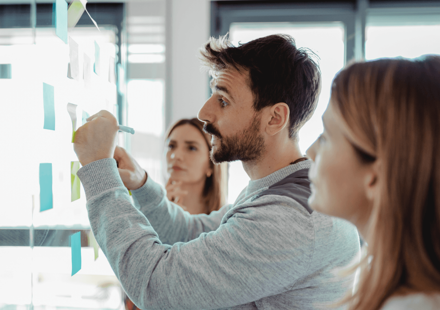 Na zdjęciu widać grupę osób współpracujących przy tablicy, do której przyklejone są kolorowe karteczki samoprzylepne (post-it). Na pierwszym planie mężczyzna w szarym swetrze pisze coś na jednej z karteczek lub nanosi poprawki, a obok niego dwie kobiety uważnie obserwują i analizują to, co się dzieje. Walidacja pomysłu na startup.